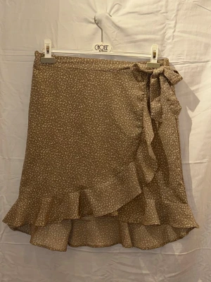 Beige omlottkjol med volang och knyt - Söt beige omlottkjol med småblommigt mönster och volangkant. Kjolen har ett knytband i midjan som ger en feminin touch. Perfekt för vår och sommar, lätt och luftig känsla.