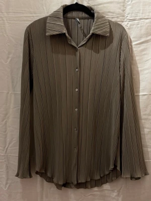 Plisserad beige skjorta med knappar - Stilren beige skjorta med plisserat tyg och klassisk krage. Skjortan har långa ärmar och knäppning framtill. Perfekt för dig som gillar en avslappnad men ändå trendig look.