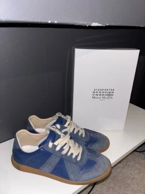 Blå Maison Margiela sneakers - Snygga blå sneakers från Maison Margiela med vita skosnören och beige gummisula. Skorna har en klassisk låg modell med ovandel i mocka och läder, perfekt för dig som gillar stilrena och trendiga sneakers.