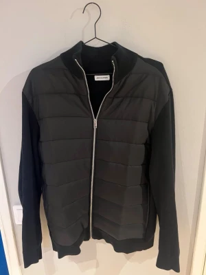 Svart Cardigan från Jack & Jones - Svart Cardigan med quiltad front och stickade ärmar. Jackan har hög ribbad krage, dragkedja framtill och ribbade muddar. Materialet är en mix av vadderad polyester och stickat tyg, vilket ger en modern look.