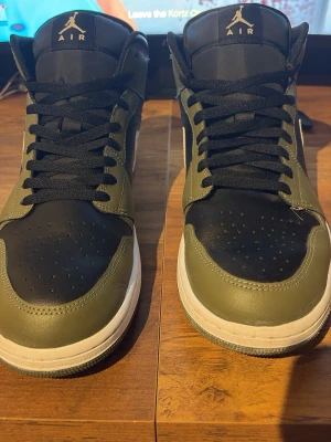 Air Jordan 1 Mid olivgrön/svart - Snygga Air Jordan 1 Mid sneakers i olivgrönt och svart läder med vita detaljer och klassisk Jumpman-logga på plösen. Skorna har svart snörning, perforerad tåbox och vit sula. Perfekta för dig som vill ha en ikonisk och stilren look.