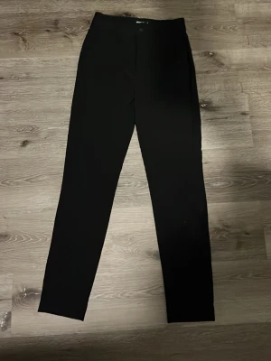 Svarta kostymbyxor med rak passform - Stilrena svarta kostymbyxor med hög midja och raka ben. Byxorna har diskreta sidofickor och stängs med knapp och dragkedja. Perfekta för en clean och klassisk look. Materialet ser ut att vara ett mjukt tyg med lite stretch.