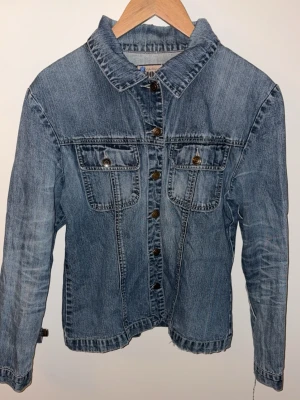 Klassisk blå jeansjacka - Snygg blå jeansjacka med raka sömmar och två bröstfickor med knappar. Jackan har en tidlös design med metallknappar framtill och på fickorna. Perfekt för dig som gillar en avslappnad och trendig stil. Materialet är slitstarkt denim.