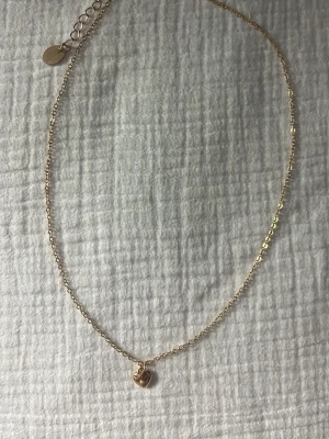 Guldhalsband med hjärtberlock - Snyggt och tunt halsband i guldfärgad metall med en liten hjärtformad berlock. Kedjan har justerbar längd med förlängningskedja. Perfekt för att addera en söt detalj till din outfit. 