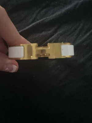 Vitt och guldigt metallarmband - Snyggt armband i vit och guldig metall med ett stort H-format spänne framtill. Armbandet har en stilren och modern design som sticker ut och passar perfekt till din outfit. Enkel att öppna och stänga tack vare det smarta spännet.