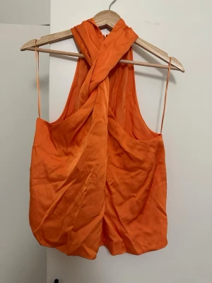 Orange omlottklänning Stockh lm Studio - Säljer en snygg orange omlottklänning från Stockh lm Studio i storlek L. Klänningen har en unik twistad halterneck och är ärmlös med kort längd. Tillverkad i ett mjukt, glansigt tyg som ger en lyxig känsla. Perfekt för dig som vill sticka ut!