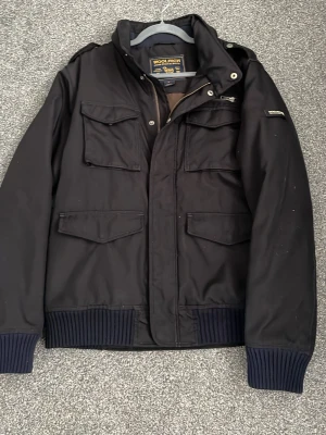 Woolrich field jacka - Snygg svart fieldjacka från Woolrich med ribbade muddar vid ärmslut och nederkant. Jackan har flera praktiska fickor framtill, axelklaffar och dragkedja med knappslå. Perfekt för dig som vill ha en stilren och funktionell jacka.