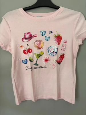 Ljusrosa t-shirt med färgglada tryck - Säljer en ljusrosa t-shirt med coola och färgglada tryck som cowboyhatt, körsbär, jordgubbe, snäcka, discokula och drink. T-shirten har korta ärmar och är gjord i mjuk bomull. Perfekt för dig som gillar lekfulla detaljer och unik stil.