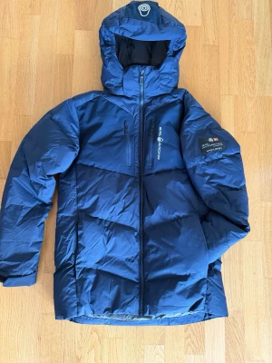 Sail Racing Jr Patrol Down Jacket - Säljer en marinblå dunjacka från Sail Racing med avtagbar huva och dragkedja framtill. Jackan har logga på bröstet och ärmen samt en bröstficka med dragkedja. Perfekt för kalla vinterdagar och riktigt snygg design.