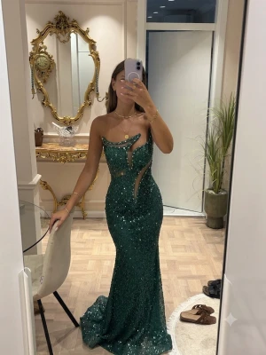 Grön glittrig långklänning - Säljer en grön glittrig långklänning med figurnära passform och unika cut-outs framtill. Klänningen är axelbandslös och har en elegant, festlig look med paljettdetaljer som verkligen sticker ut. Jag är 170cm lång och klänningen är specialsydd till mina mått.   Klänningen är köpt från Zana Fashion Group https://www.instagram.com/zanafashiongroup?igsh=NnA5azFtMW81a2E=