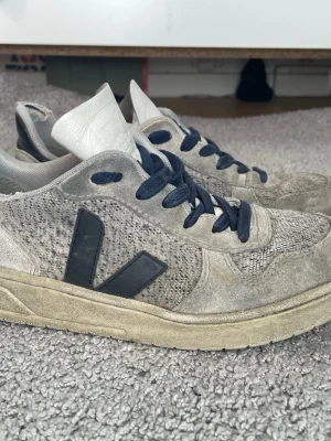 Veja sneakers med mockadetaljer - Säljer ett par gråa Veja sneakers med ovandel i mocka och textil, svarta detaljer och snörning. Skorna har en tydlig Veja-logga på sidan och svart hälparti med logotyp. Snygg retrostil och passar perfekt till streetwear för killar och tjejer. Storlek = 38