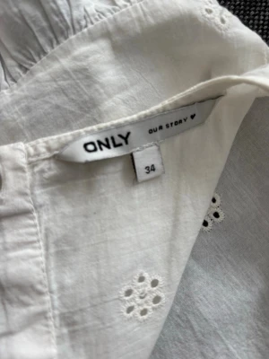 Vit broderad blus från ONLY - Söt vit blus från ONLY i storlek 34 med broderade blommönster och volangdetaljer vid ärmhålen. Blusen är ärmlös och har knappar längs ryggen samt en vågig kant nedtill. Tillverkad i ett lätt bomullstyg som känns luftigt och somrigt.