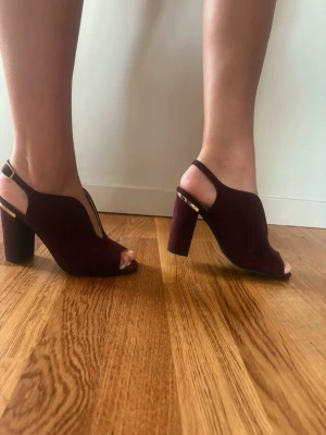 Vinröda chunky pumps med öppen tå - Säljer ett par vinröda pumps med chunky klack och öppen tå. Skorna har en snygg rem med gulddetalj runt hälen och är gjorda i mockaimitation. Perfekta för dig som vill ha en statement-sko med modern vibe.