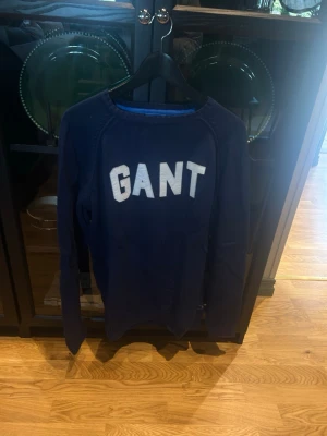 Mörkblå GANT tröja med logga - Snygg mörkblå tröja från GANT med stor vit logga på bröstet. Klassisk rund halsringning och långa ärmar. Perfekt för dig som gillar stilrena och sportiga plagg. Tillverkad i mjukt material för skön känsla. Pruta!!