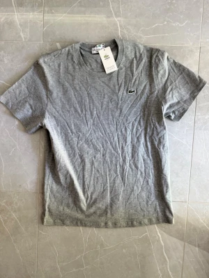 Grå t-shirt från Lacoste - Snygg grå t-shirt från Lacoste med klassisk rund hals och det ikoniska krokodilmärket broderat på bröstet. Tillverkad i mjuk bomull och har en enkel, stilren design som passar till det mesta.