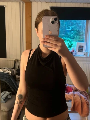 Svart halterneck topp med öppen rygg - Snygg svart halterneck topp med öppen rygg och smal passform. Toppen är ärmlös och har en cool detalj med band runt nacken. Perfekt för dig som gillar stilrena och trendiga plagg.