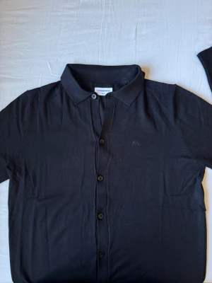 Svart skjorta med broderad logga - Stilren svart skjorta med klassisk krage och knappar framtill. Skjortan har en diskret broderad logga på bröstet och är tillverkad i ett mjukt bomullstyg. Perfekt för dig som gillar enkel och clean stil.