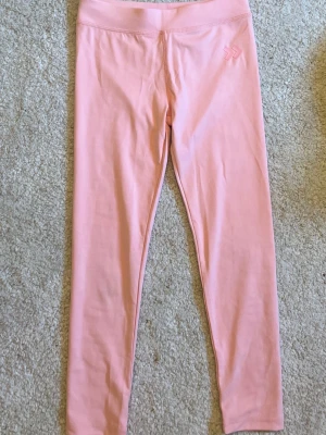 Ljusrosa leggings - Säljer ett par ljusrosa leggings med diskret logga på höften. De har hög midja och är tillverkade i ett mjukt, stretchigt material som sitter skönt mot kroppen. Perfekta för en avslappnad och trendig look.