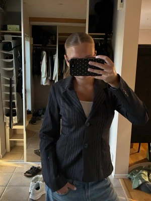 Mörkgrå kavaj med ränder från Vila - Snygg mörkgrå kavaj från Vila med tunna vertikala ränder i beige. Figurnära passform, två knappar framtill och klassisk krage. Fodrad insida för extra komfort. Perfekt att styla med jeans eller kjol för en trendig look.