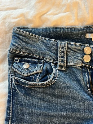 Blå jeans med dubbla knappar - Säljer ett par blå jeans med snygg tvätt, dubbla knappar i midjan och coola detaljer på fickorna. Jeansen har raka ben och klassisk femficksdesign. Materialet är denim i bomull med kontrastsömmar för en trendig look.