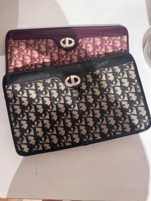 Svart och beige monogram kuvertväska - Säljer en snygg kuvertväska i svart och beige med klassiskt monogrammönster och silverfärgat vridlås. Väskan har detaljer i svart läder och en stilren, rektangulär form. Perfekt för dig som gillar tidlös design och vill ha något som sticker ut.