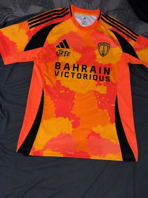 Orange Paris FC matchtröja - Säljer en färgstark matchtröja från Paris FC med digitalt pixelmönster i orange, rött och gult. Tröjan har svarta detaljer, korta ärmar och tryck med 'BAHRAIN VICTORIOUS' på bröstet. Perfekt för fotboll eller träning.