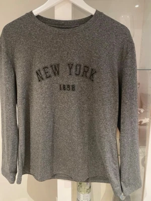Grå sweatshirt med New York-tryck - Grå långärmad sweatshirt med texten 'NEW YORK 1898' tryckt på bröstet. Tröjan har rund halsringning och är gjord i mjukt sweatshirtmaterial. Perfekt för en avslappnad och trendig stil.