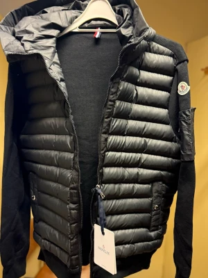 Svart pufferjacka från Moncler - Snygg svart pufferjacka från Moncler med quiltad front och huva. Jackan har stickade ärmar och rygg, dragkedja framtill och fickor med tryckknappar. Moncler-logga på ärmen och en liten ficka på vänster arm. Perfekt för kalla vinterdagar.