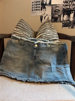 Blå jeanskjol Nelly  - Trendig blå jeanskjol i kort modell med råa, fransiga kanter både upptill och nertill. Klassisk femficksdesign och knappgylf framtill. Perfekt för dig som gillar en avslappnad och edgy stil.