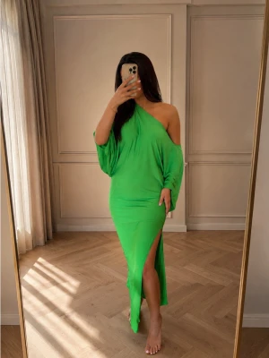 Grön oneshoulder långklänning - Snygg grön långklänning med oneshoulder-design och slits på sidan. Klänningen har en lös passform upptill och är figurnära nertill. Perfekt för dig som vill sticka ut med en färgstark look.