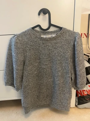 Grå tröja med puffärm från 157 - Mysig grå tröja från 157 med puffärm och rund halsringning. Tröjan har en mjuk, borstad yta och trekvartsärmar som ger en trendig look. Perfekt att matcha med jeans eller kjol för en avslappnad stil.