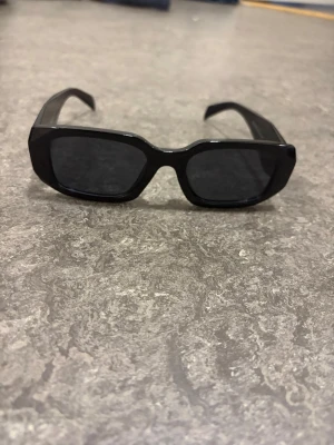 Prada solglasögon  - Snygga svarta solglasögon från Prada med rektangulära bågar och mörka glas. Klassisk och stilren design med breda skalmar där Prada-loggan syns tydligt på sidan. Perfekta för dig som vill ha en trendig accessoar till sommaren.