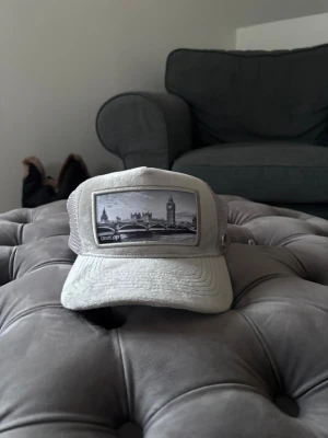 Grå keps med London-motiv - Snygg grå keps med mesh på sidorna och ett stort tygpatch framtill med svartvitt motiv av London och Big Ben. Justerbar baktill för perfekt passform. Perfekt accessoar för dig som gillar streetstyle och vill sticka ut med en unik detalj.