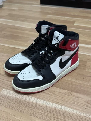 Nike Air Jordan 1 High svart/vit/röd - Säljer ett par Nike Air Jordan 1 High sneakers i svart, vitt och rött. Klassisk high-top siluett med svart swoosh, perforerad tåbox och röda detaljer vid hälen och kragen. Skorna har vita paneler och svart snörning. Perfekta för dig som gillar streetwear.