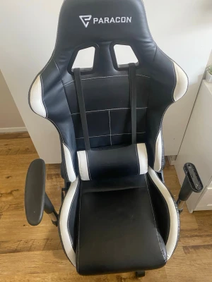 Gamingstol Paracon - Säljer en svart och vit gamingstol från Paracon med hög rygg, justerbara armstöd och extra nackkudde. Stolen har sportig design med vita detaljer längs kanterna och är klädd i konstläder. Perfekt för långa spelsessioner eller hemmakontoret. Armstöd är inte fast skruvad. Det lösa armstödet medföljer, så att man kan sätta fast det igen!