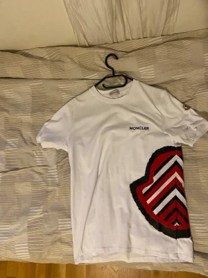  Moncler Vit t-shirt med logga - Snygg vit t-shirt från moncler  med klassisk rund halsringning och broderad logga på ärmen. Tillverkad i mjuk bomull som känns skön mot huden. Perfekt för en clean och stilren look.