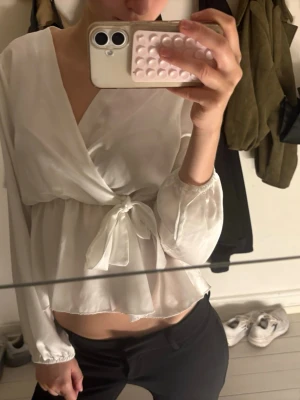 Vit omlottblus med knytband - Snygg vit omlottblus med långa ärmar och knytband i midjan. Blusen har v-ringning och är gjord i ett lätt, glansigt material som ger en elegant look. Perfekt att styla med både jeans och kjol för en trendig outfit.