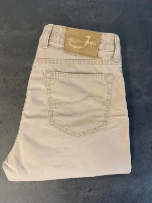 Beige jeans från Jacob Cohen, stl 32 - Snygga beige jeans från Jacob Cohen med klassisk femficksmodell och raka ben. Tillverkade i mjukt bomullstyg med diskreta sömmar och patch med logga bak i midjan. Perfekta för en clean och stilren look.