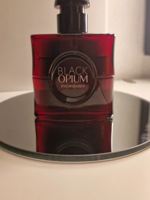 Yves Saint Laurent Black Opium Red - Black Opium Eau de Parfum Over Red från Yves Saint Laurent. Flaskan är djup röd med svart rund etikett och elegant svart lock. Parfymen kommer i originalförpackning och har en lyxig, modern design som sticker ut på hyllan.