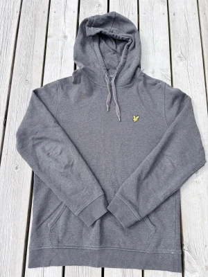 Grå hoodie från Lyle & Scott - Snygg grå hoodie från Lyle & Scott med klassisk gul logga på bröstet. Hoodien har huva med dragsko, magficka och ribbade muddar vid ärmslut och nederkant. Tillverkad i mjukt bomullsmaterial som är skönt att bära.