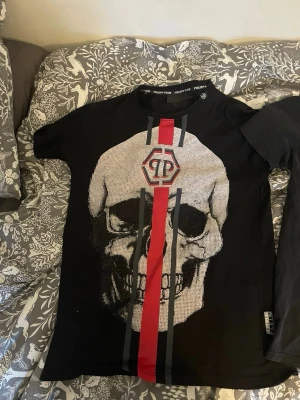 Svart t-shirt med döskalle och ränder - Säljer en svart t-shirt med stor döskalle framtill, röda och grå ränder samt PP-logga. Baksidan har döskalletryck och röda detaljer. T-shirten har rund hals och korta ärmar, perfekt för dig som gillar edgy streetwear.