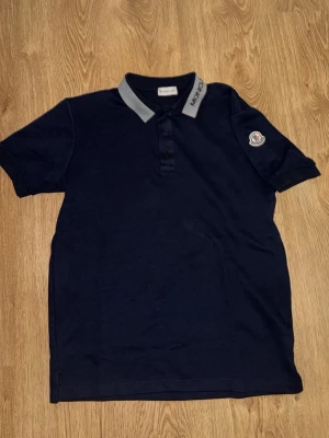 Mörkblå Moncler pikétröja - Snygg mörkblå pikétröja från Moncler med grå krage och logga på ärmen. Klassisk pikéknäppning framtill och diskret Moncler-tryck på kragen. Perfekt till jeans eller shorts för en clean look.