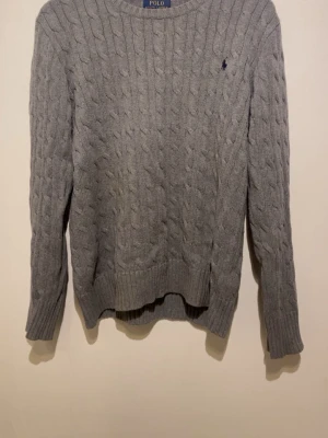 Grå kabelstickad tröja Polo Ralph Lauren - Kabelstickad grå tröja från Polo Ralph Lauren med rund halsringning och klassisk broderad logga på bröstet. Långa ärmar och ribbade muddar ger en snygg och tidlös look. Perfekt för lager-på-lager-stil. Priset är inte hugget i sten, släng iväg ett dm om du har några frågor 😁