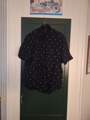 Svart kortärmad skjorta med palmer - Snygg svart skjorta med korta ärmar och små vita palmträd som mönster. Perfekt för en avslappnad och cool stil. Skjortan har knappar framtill och klassisk krage.