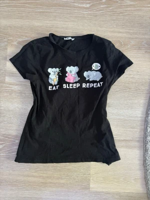 Svart t-shirt med koalamotiv från Siste - Cool svart t-shirt från New Yorker med tryck av tre koalor och texten 'EAT SLEEP REPEAT' på framsidan I storlek L, kan användas som tröja eller nattlinne om man är yngre. Inga hål eller synliga fläckar.