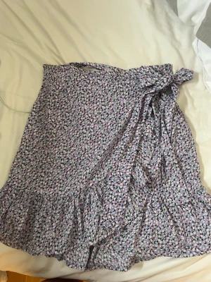 Ljuslila blommig omlottkjol med volang - Supersöt ljuslila omlottkjol med små blommor i svart, vit och grön. Kjolen har en knytning i midjan och volangkant nedtill som ger en extra feminin touch. Perfekt för vår och sommar!