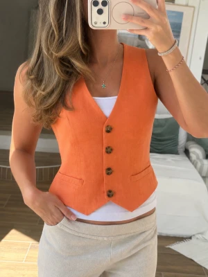 Orange linneväst med knappar - Snygg orange väst i linne med fyra bruna knappar framtill och två fickdetaljer. Västen har en v-ringad skärning och en figurnära passform som ger en trendig look. Perfekt att styla över en t-shirt eller skjorta för en cool och modern outfit.