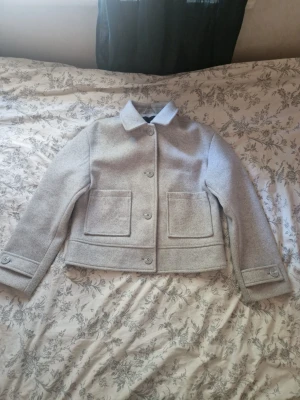 Ljusgrå kappa med stora fickor - Snygg ljusgrå jacka med stora framfickor och klassisk krage. Jackan har raka linjer, stora knappar framtill och är helfodrad. Perfekt för dig som gillar stilrena och minimalistiska plagg.