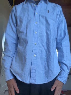 Ljusblå skjorta med knappstängning - Klassisk ljusblå skjorta med långa ärmar och knappar framtill. Skjortan har en liten broderad logga på bröstet och en normal passform. Perfekt till jeans eller chinos för en clean look.