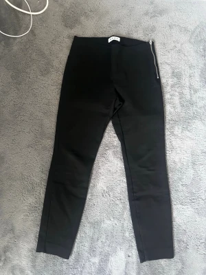 Svarta leggings med dragkedja - Säljer ett par svarta leggings med smal passform och hög midja. De har en snygg dragkedja i sidan som detalj och är gjorda i ett stretchigt material som sitter skönt på kroppen. Perfekta till en avslappnad eller trendig look.
